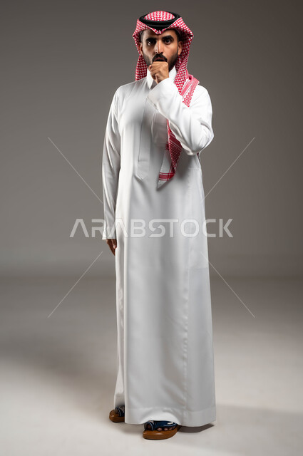 الشعور بالإرهاق والتعب، الإصابة بالأنفلونزا ونزلات البرد، الكحة والتهاب الحلق والحاجة للعلاج، بورتريه مقرب لرجل عربي سعودي خليجي يرتدي الشماغ والثوب التقليدي ينظر إلى الكاميرا يضع يده أمام فمه ويقوم بالسعال, صورة الجسم بالطول الكامل، خلفية رمادية .
