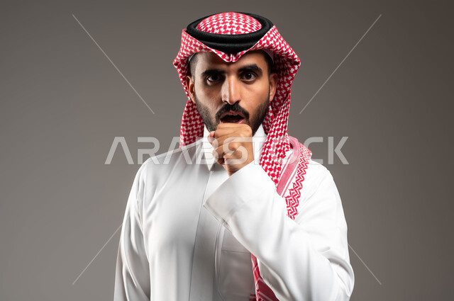 الكحة والتهاب الحلق والحاجة للعلاج، بورتريه مقرب لرجل عربي سعودي خليجي يرتدي الشماغ والثوب التقليدي ينظر إلى الكاميرا يضع يده أمام فمه ويقوم بالسعال, الإصابة بالأنفلونزا ونزلات البرد، خلفية رمادية .