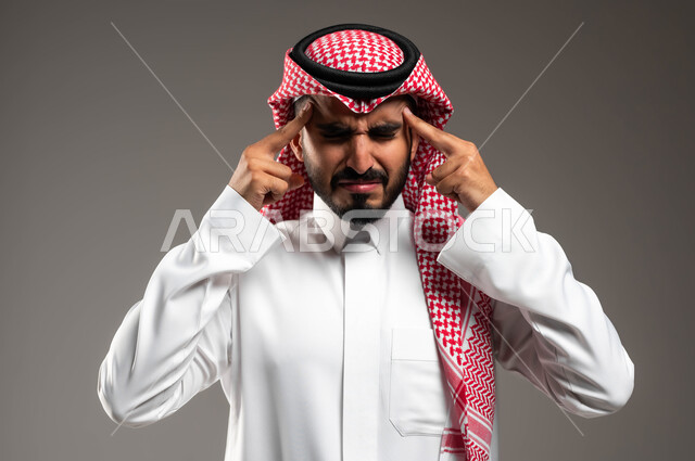 The need to seek help and treatment, suffering from migraine attacks, headache gestures and feeling sick, a close-up portrait of a Saudi Gulf Arab man wearing a shemagh and traditional dress, holding his head in his hands out of intense pain, feeling tired and exhausted, gray background