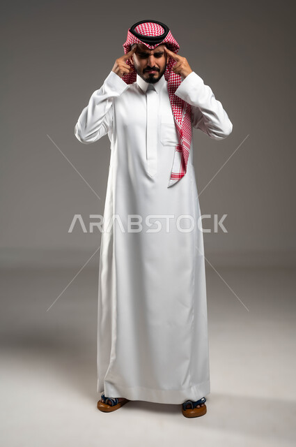 الإصابة بنوبات الشقيقة، إيماءات الصداع والإحساس بالمرض, الحاجه للعلاج وطلب المساعده, بورتريه لشاب عربي سعودي خليجي يرتدي الشماغ والثوب التقليدي يمسك رأسه بيديه من شدة الألم, الشعور بالتعب والإرهاق, صورة الجسم بالطول الكامل، خلفية رماديه .