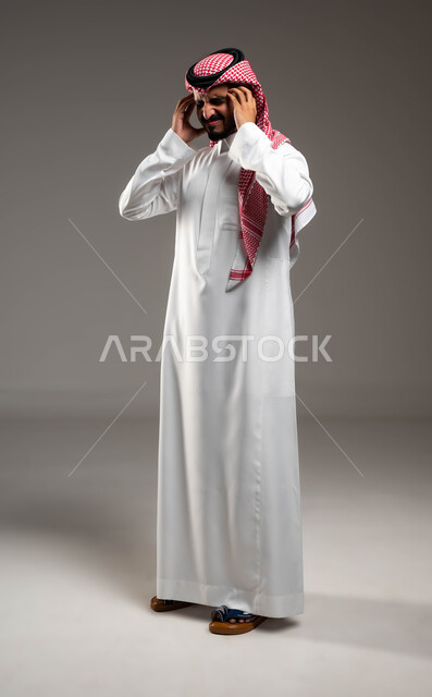 إيماءات الصداع والإحساس بالمرض, الحاجه للعلاج وطلب المساعده, بورتريه لشاب عربي سعودي خليجي يرتدي الشماغ والثوب التقليدي يمسك رأسه بيديه من شدة الألم, الشعور بالتعب والإرهاق, الإصابة بنوبات الشقيقة، صورة الجسم بالطول الكامل، خلفية رماديه .