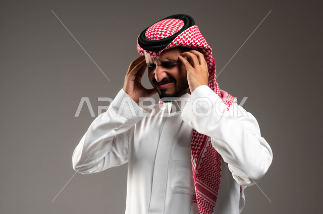 الحاجه للعلاج وطلب المساعده, بورتريه مقرب لشاب عربي سعودي خليجي يرتدي الشماغ والثوب التقليدي يمسك رأسه بيديه من شدة الألم, إيماءات الصداع والإحساس بالمرض, الشعور بالتعب والإرهاق, الإصابة بنوبات الشقيقة، خلفية رماديه .