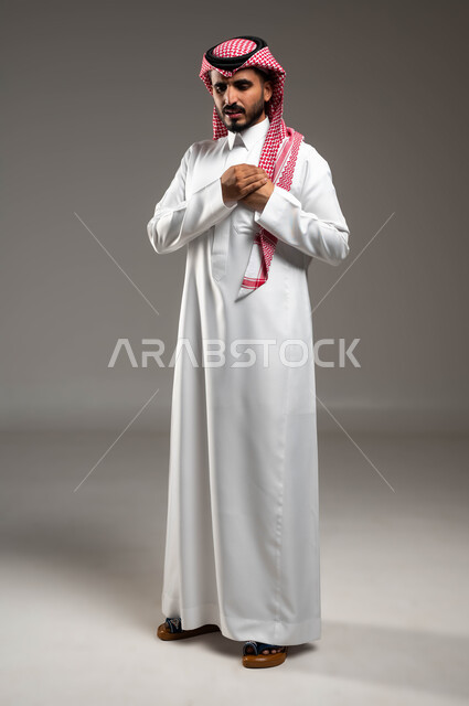 الذبحه الصدريه حالة مرضية، إيماءات التعب والإرهاق والضغوطات النفسية، بورتريه لشاب عربي سعودي خليجي يرتدي الشماغ و الثوب التقليدي يضع يده على قلبه يشعر بحالة سيئة من الوخز والألم في قلبه, صورة الجسم بالطول الكامل، خلفية رمادية .