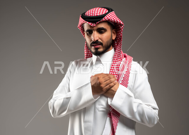 مشاكل صحية صعبة، الذبحه الصدريه حالة مرضية، إيماءات التعب والإرهاق والضغوطات النفسية، بورتريه مقرب لشاب عربي سعودي خليجي يرتدي الشماغ و الثوب التقليدي يضع يده على قلبه يشعر بحالة سيئة من الوخز والألم في قلبه,  خلفية رمادية .