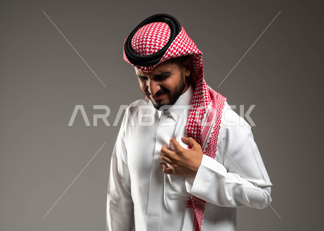 أزمات قلبية مفاجئة، الذبحه الصدريه حالة مرضية، إيماءات التعب والإرهاق والضغوطات النفسية، بورتريه لشاب عربي سعودي خليجي يرتدي الشماغ و الثوب التقليدي يضع يده على قلبه يشعر بحالة سيئة من الوخز والألم في قلبه,  خلفية رمادية .