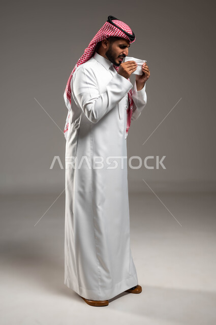 ايماءات العطاس والإرهاق، استخدام المناديل الورقية, الإصابة بالزكام والانفلونزا، بورتريه لرجل عربي سعودي خليجي يرتدي الشماغ و الثوب التقليدي يشعر بالمرض والتعب الشديد, صورة الجسم بالطول الكامل، خلفية رمادية .