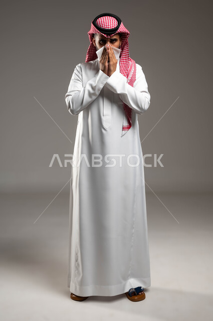 إيماءات المرض والتعب الشديد، تعابير تدل على العطاس والإرهاق، الإصابة بالزكام والانفلونزا، بورتريه لرجل عربي سعودي خليجي يرتدي الشماغ و الثوب التقليدي يمسح أنفه باستخدام المناديل الورقية, صورة الجسم بالطول الكامل، خلفية رمادية .