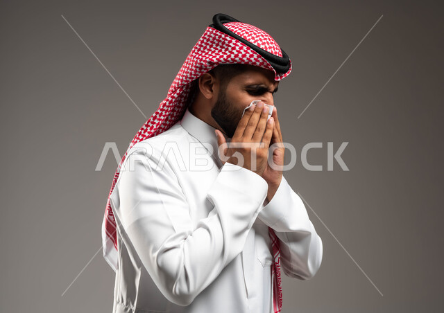 الإصابة بالزكام والانفلونزا، بورتريه مقرب لرجل عربي سعودي خليجي يرتدي الشماغ و الثوب التقليدي يمسح أنفه باستخدام المناديل الورقية, إيماءات تعبر عن المرض والتعب الشديد، اشارات تدل على العطاس والإرهاق، خلفية رمادية .