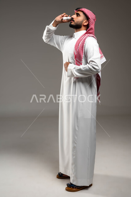 إيماءات المرض والتعب الشديد، الإصابة بالزكام والانفلونزا، بورتريه لرجل عربي سعودي خليجي يرتدي الشماغ و الثوب التقليدي يستخدم بخاخة الأنف, المعاناة من الحساسية المزمنة و التهاب الجيوب الأنفية, صورة الجسم بالكامل، خلفية رمادية
