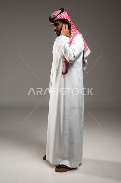 الحاجة للمساعدة و للذهاب للطبيب و وضع خطة علاجية، التهاب الأذن الوسطى، بورتريه من الجانب لشاب عربي سعودي خليجي مريض يرتدي الشماغ و الثوب التقليدي السعودي يضع يده على اذنه بايماءات التألم الشديد، صورة الجسم بالطول الكامل، خلفية رمادية
