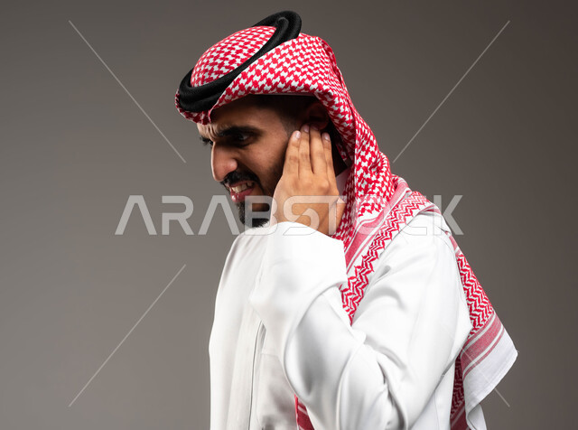 التهاب الأذن الوسطى، الحاجة للذهاب للطبيب للمساعدة و وضع خطة علاجية مناسبة، بورتريه مقرب من الجانب لشاب عربي سعودي خليجي يرتدي الشماغ و الثوب التقليدي السعودي يضع يده على اذنه بايماءات التألم الشديد، حالة اسعافية طارئة، خلفية رمادية