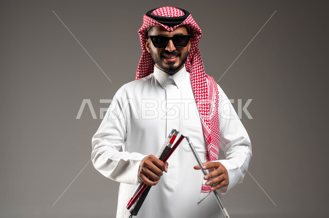 تأمين المستلزمات الآمنة الخاصة بالمكفوفين  لتحقيق رغباتهم، بورتريه مقرب لرجل عربي سعودي خليجي ضرير مبتسم يرتدي الشماغ والثوب التقليدي يضع النظارة الشمسية يقوم بتجهيز العصا المساعدة في التوازن و عدم التعثر، ايماءات الثقة بالنفس و الرضا، خلفية رمادية