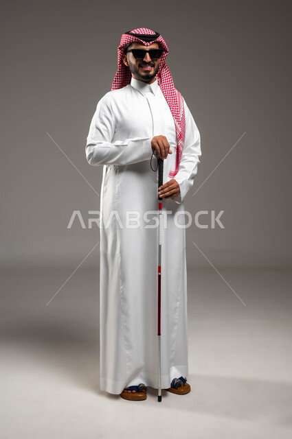 Using a cane that helps with balance and mobility without stumbling, a portrait of a blind Arab Saudi Gulf man wearing sunglasses holding a cane, providing safe roads for the blind in Saudi Arabia, tools and equipment that help blind people, full-length body portrait, gray background