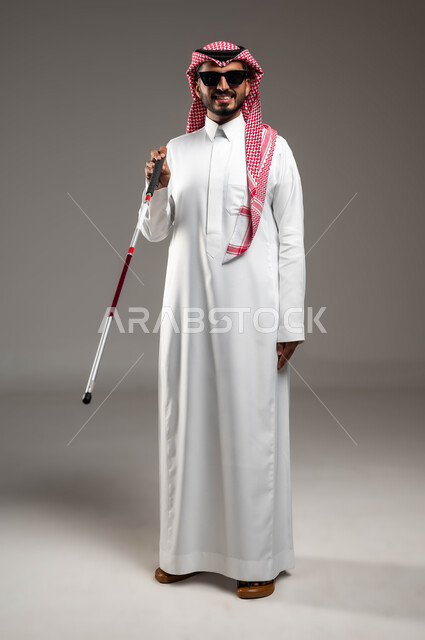 Teaching the blind about mobility techniques using a cane, a portrait of a blind Saudi Arabian Gulf man wearing a shemagh and traditional dress, smiling, wearing sunglasses, holding a white cane, providing psychological support to the blind to enhance their confidence and motivate them to achieve their potential, full-body portrait, gray background