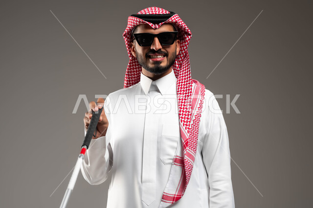 الاعتماد على الدلائل الصوتية المحيطة، التحسس للعقبات وتجنبها أثناء المشي، بورتريه مقرب لرجل عربي سعودي خليجي ضرير مبتسم يرتدي الشماغ والثوب التقليدي يضع النظارة الشمسية يمسك العصا لمساعدته في التنقل، ايماءات الثقة بالنفس، خلفية رمادية
