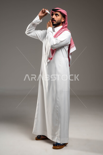 الاحساس بالحرقة و احمرار العيون، الحاجة للفحص و علاج حالات التهاب العصب البصري، بورتريه من الجانب لرجل عربي خليجي سعودي مريض يرتدي الشماغ والثوب التقليدي يقوم بفتح عينه و وضع القطرة فيها، صورة الجسم بالطول الكامل، ارتفاع ضغط العين، خلفية رمادية