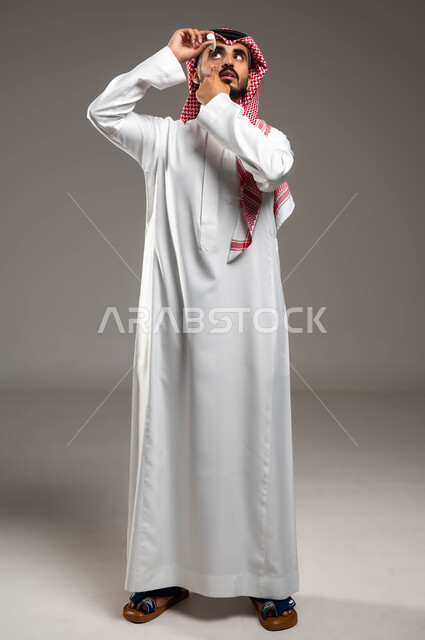 الاحساس بالحرقة و احمرار العيون، الحاجة للفحص و علاج حالات التهاب العصب البصري، بورتريه لرجل عربي خليجي سعودي مريض يرتدي الشماغ والثوب التقليدي يقوم بوضع قطرة العين، صورة الجسم بالطول الكامل، ارتفاع ضغط العين، خلفية رمادية