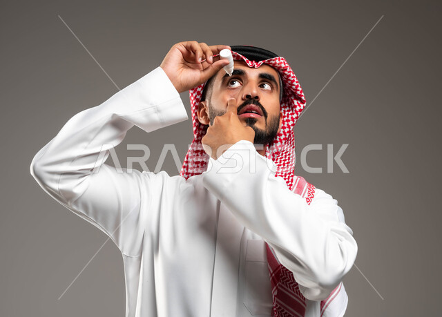 مشاكل صحية و حالات مرضية، التهاب العصب البصري، بورتريه مقرب لرجل عربي خليجي سعودي مريض يرتدي الشماغ والثوب التقليدي يقوم بوضع قطرة العين، الحاجة للفحص والعلاج، التهاب القرنية العينية، ايماءات الحرقة و وجع و احمرار العيون، خلفية رمادية