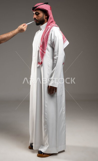الحاجة إلى العلاج والمساعده, الإحساس والشعور بالمرض والإرهاق، بورتريه لشاب عربي سعودي خليجي يرتدي الشماغ والثوب التقليدي يعاني من حمى شديدة، قياس درجة الحرارة بإستخدام الميزان الرقمي، صورة الجسم بالكامل، خلفية رمادية