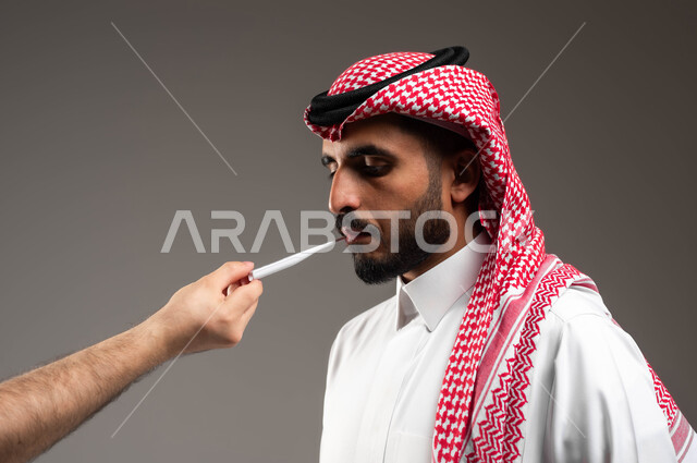 Measuring temperature using a digital thermometer, the need for treatment and assistance, a close-up portrait from the side of a young Saudi Arabian Gulf man wearing a shemagh and traditional dress, suffering from a high fever, feeling sick and tired, gray background.