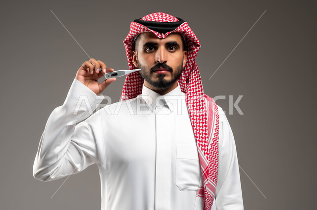 أهمية وجود جهاز مقياس حرارة في المنزل، بورتريه مقرب لرجل عربي سعودي خليجي يرتدي الشماغ والثوب التقليدي ينظر الى الكاميرا بإيماءات الحزن واليأس بسبب المرض، القيام بعرض الميزان الرقمي، الشعور بالتعب والإرهاق، خلفية رمادية