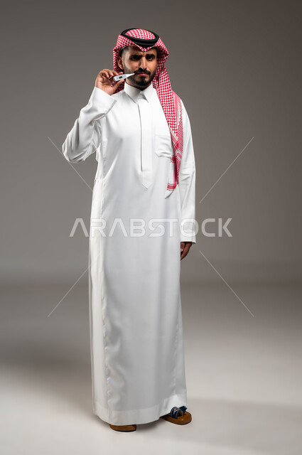 النظر الى الكاميرا بإيماءات التعب والإرهاق بسبب الإصابة بالحمى، بورتريه لرجل عربي سعودي خليجي يرتدي الشماغ والثوب التقليدي يقوم بقياس حرارته بإستخدام جهاز قياس درجة الحرارة، صورة الجسم بالكامل، خلفية رمادية