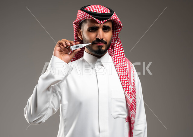 ضرورة زيارة الطبيب لوضع الخطة العلاجية، الإحساس بالمرض والإرهاق، بورتريه مقرب لشاب عربي سعودي خليجي يرتدي الشماغ والثوب التقليدي يقوم بقياس درجة حرارته بإستخدام الميزان الرقمي ينظر الى الكاميرا بتعابير الانزعاج والتعب، خلفية رمادية