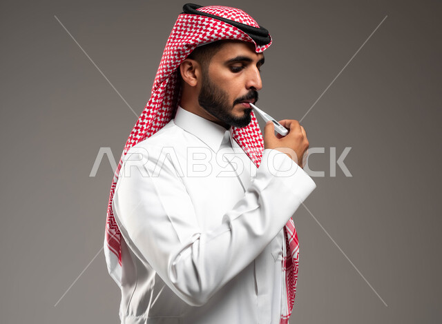 الإحساس بالمرض والإرهاق، الإصابة بالحمى والتعب، بورتريه مقرب من الجانب لشاب عربي سعودي خليجي يرتدي الشماغ والثوب التقليدي يقوم بقياس حرارته باستخدام الميزان الرقمي، الحاجة إلى زيارة الطبيب للمساعدة ووضع خطة علاجية, خلفية رمادية