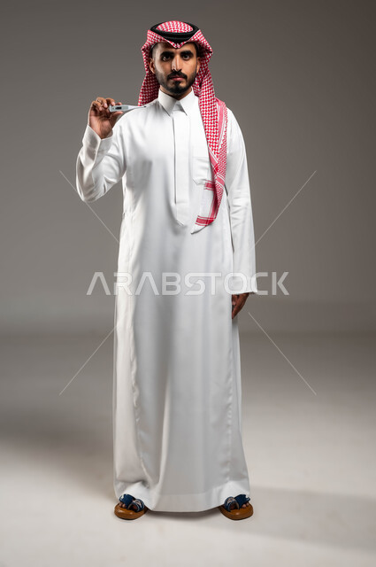 ارتفاع درجات حرارة الجسم والشعور بالتعب والإرهاق، بورتريه لرجل عربي سعودي خليجي يرتدي الشماغ والثوب التقليدي يمسك بيده جهاز مقياس الحرارة، النظر الى الكاميرا بإيماءات اليأس والحزن, صورة الجسم بالكامل، خلفية رمادية