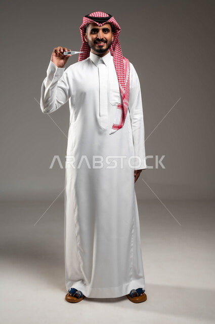 الشعور بالتحسن وانخفاض الحرارة، مفهوم التماثل للشفاء، بورتريه لشاب عربي خليجي سعودي مبتسم يرتدي الشماغ والثوب التقليدي يحمل بيده جهاز مقياس الحرارة، النظر الى الكاميرا بتعابير السرور والفرح للتعافي من المرض، صورة الجسم بالطول الكامل، خلفية رمادية