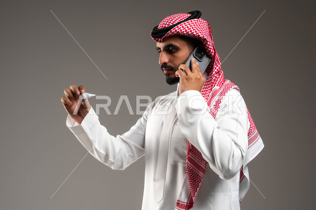 اجراء مكالمة هاتفية عبر الجوال لاستشارة الطبيب عن العلاج المناسب، الشعور بالتعب والإرهاق وأعراض الحمى، بورتريه مقرب لشاب عربي سعودي خليجي يرتدي الشماغ والثوب التقليدي يمسك بيده جهاز مقياس الحرارة ينظر الى المؤشر، خلفية رمادية