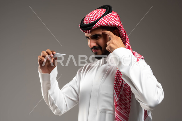 الاصابة بالصداع ونوبات الشقيقة، بورتريه مقرب لرجل عربي سعودي خليجي يرتدي الشماغ والثوب التقليدي يمسك جهاز مقياس الحرارة يضع يده على رأسه من شدة الألم, الشعور بالتعب والإرهاق و ارتفاع درجات حرارة الجسم، خلفية رمادية