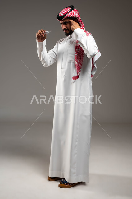 الشعور بالتعب والإرهاق وأعراض الحمى، إيماءات الصداع والإصابة بنوبات الشقيقة، بورتريه لشاب عربي سعودي خليجي يرتدي الشماغ والثوب التقليدي يمسك جهاز مقياس الحرارة يضع يده على رأسه من شدة الألم, صورة الجسم بالكامل، خلفية رمادية