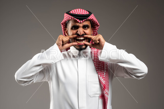 النظر إلى الكاميرا بتعابير الخوف, بورتريه مقرب لرجل عربي خليجي سعودي يرتدي الشماغ والثوب التقليدي يقوم بفتح فمه بشكل مخيف، الحاجة للزيارة الدورية الى طبيب الاسنان للتخلص من الألام، خلفية رمادية