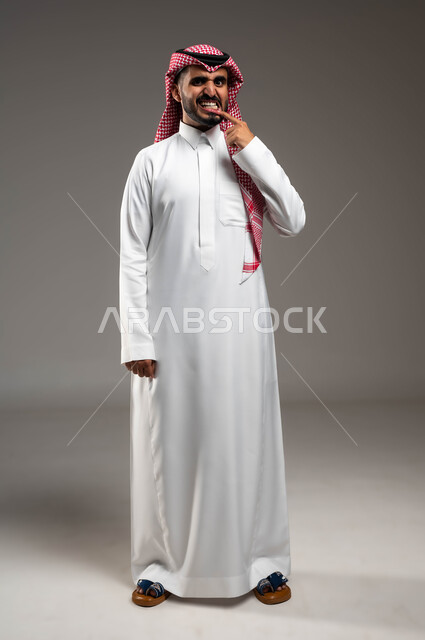 إبتسامة هوليود باستخدام قشور فينير من الخزف، بورتريه لرجل عربي خليجي سعودي مبتسم يرتدي الشماغ والثوب التقليدي ينظر إلى الكاميرا يشير بإصبعه إلى أسنانه، الاهتمام والعناية بالصحة الفموية، أهمية الزيارة الدورية إلى طبيب الأسنان، صورة لكامل الجسم، خلفية رمادية