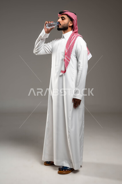 الحفاظ على سلامة الجهاز المناعي، المحافظة على صحة الكلى ومعالجتها، بورتريه لرجل عربي خليجي سعودي يرتدي الشماغ والثوب التقليدي يقوب بشرب المياه من عبوة بلاستيكية، أهمية شرب الماء النقي للمحافظة على بشرة نضرة، التخلص من سموم الجسم، صورة الجسم بالكامل، خلفية رمادية
