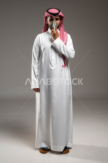 استنشاق أدوية معالجة لانقطاع التنفس، المحافظة على ممر هوائي منتظم باستخدام جهاز التنفس الصناعي، بورتريه لرجل عربي سعودي خليجي يرتدي الشماغ والثوب التقليدي يقوم بوضع قناع الاكسجين، مداواة أمراض الربو المزمنة، صورة الجسم بالطول الكامل، خلفية رمادية