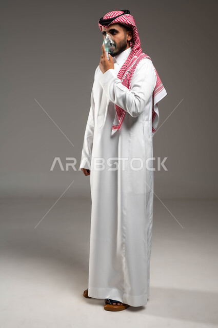 الحفاظ على ممر هوائي منتظم باستخدام جهاز التنفس الصناعي، بورتريه لرجل عربي سعودي خليجي يرتدي الشماغ والثوب التقليدي يقوم بوضع قناع الاكسجين، مداواة أمراض الربو المزمنة، استنشاق أدوية معالجة لانقطاع التنفس، صورة الجسم بالطول الكامل، خلفية رمادية