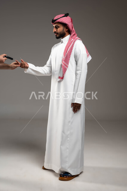 التشخيص السريع والفحص الطبي المنزلي، بورتريه من الجانب لشاب عربي خليجي سعودي مريض يرتدي الشماغ والثوب التقليدي يقوم بقياس نسبة السكر في دمه باستخدام جهاز إلكتروني حديث لمرضى داء السكري، صورة لكامل الجسم، خلفية رمادية
