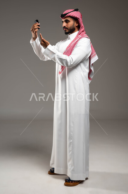 التشخيص السريع والفحص الطبي المنزلي، بورتريه من الجانب لشاب عربي خليجي سعودي مريض يرتدي الشماغ والثوب التقليدي يقوم بقياس نسبة السكر في دمه باستخدام جهاز إلكتروني حديث لمرضى داء السكري، صورة لكامل الجسم، خلفية رمادية