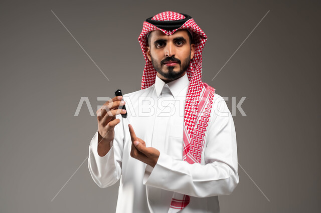 النظر الى الكاميرا بتعابير اليآس والتعب، بورتريه مقرب لشاب عربي خليجي سعودي مريض يرتدي الشماغ والثوب التقليدي يقوم بفحص نسبة السكر في دمه باستخدام جهاز منزلي الكتروني حديث لمرضى داء السكري، خلفية رمادية