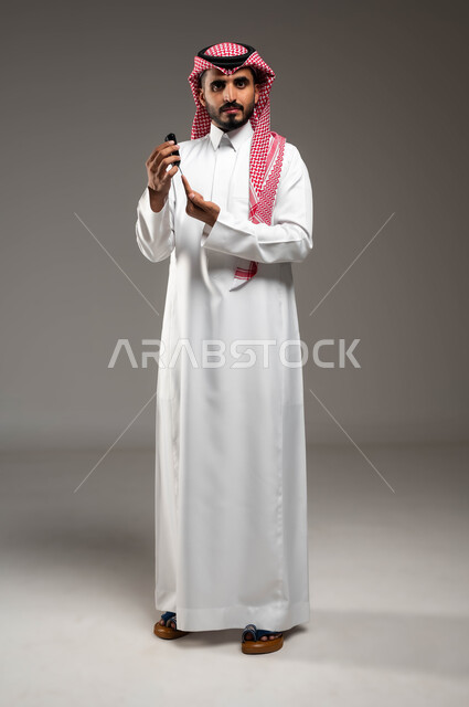 A modern electronic device to measure blood sugar for diabetics, a close-up portrait of a sick young Saudi Gulf Arab man wearing a shemagh and traditional dress, checking his blood sugar level, looking at the camera with expressions of despair and fatigue, full body image, problems with high blood sugar, gray background