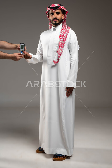 مشاكل وآلام صحية، جهاز إلكتروني حديث لمرضى داء السكري، بورتريه لشاب عربي خليجي سعودي مريض يرتدي الشماغ والثوب التقليدي يقوم بفحص نسبة السكر في دمه ينظر الى الكاميرا بتعابير اليأس والتعب، صورة الجسم بالكامل، خلفية رمادية