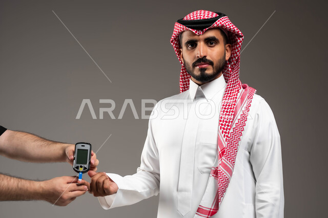 الإصابة بداء السكري، بورتريه مقرب لشاب عربي خليجي سعودي مريض يرتدي الشماغ والثوب التقليدي يقوم بفحص نسبة السكر في دمه ينظر الى الكاميرا بتعابير التعب، أجهزة الكترونية حديثة للفحص الطبي السريع، خلفية رمادية