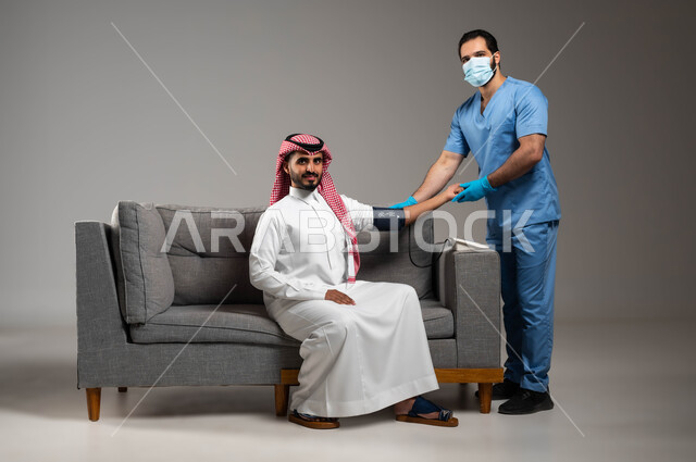 العمل في القطاع الصحي السعودي، الفحص الطبي والتشخيص السريع والرعاية الصحية المنزلية، بورتريه لممرض يرتدي الكمامة وقفازات الوقاية يقوم بقياس ضغط الدم لمريض عربي خليجي سعودي يرتدي الزي التقليدي يجلس على الكنبه، متابعة دورية لفحص ضغط الدم في البيت، خلفية رمادية