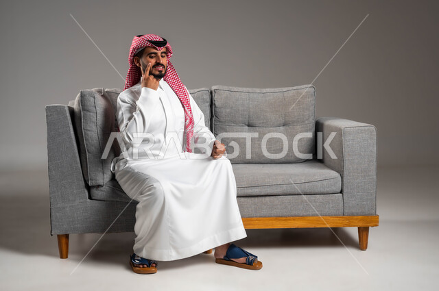 الإصابة بالحساسية الجلدية, بثور منتشرة على بشرة الوجه, الإشارة إلى مكان الإصابة, بورتريه لرجل عربي خليجي سعودي يرتدي الثوب التقليدي والشماغ يجلس على الكنبه بإيماءات الألم, المشاكل الصحية والتهجيات والشعور بالأوجاع والالام, خلفية رمادية