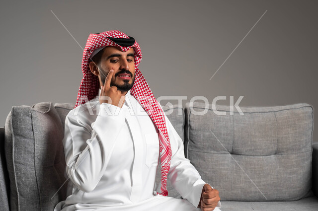 بثور منتشرة على بشرة الوجه, الإشارة إلى مكان الإصابة, بورتريه مقرب لرجل عربي خليجي سعودي يرتدي الثوب التقليدي والشماغ يجلس على الكنبه بإيماءات الاستغراب والألم, الإصابة بالحساسية الجلدية, المشاكل الصحية والتهجيات والشعور بالأوجاع والالام, خلفية رمادية