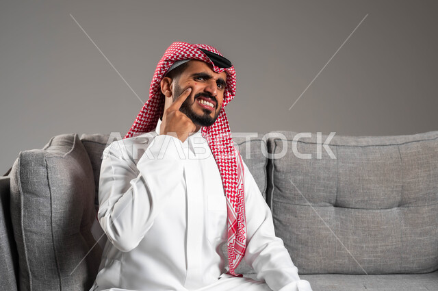 المشاكل الصحية والتهجيات والشعور بالأوجاع والالام, الإشارة إلى مكان الإصابة, بثور منتشرة على بشرة الوجه, بورتريه مقرب لرجل عربي خليجي سعودي يرتدي الثوب التقليدي والشماغ يجلس على الكنبه بإيماءات الإنزعاج والتعب, الإصابة بالحساسية الجلدية, خلفية رمادية