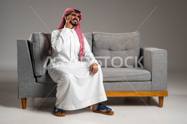 الإشارة إلى مكان الإصابة, بثور منتشرة على بشرة الوجه, بورتريه لرجل عربي خليجي سعودي يرتدي الثوب التقليدي والشماغ يجلس على الكنبه بإيماءات الإنزعاج والتعب, الإصابة بالحساسية الجلدية, المشاكل الصحية والتهجيات والشعور بالأوجاع والالام, خلفية رمادية