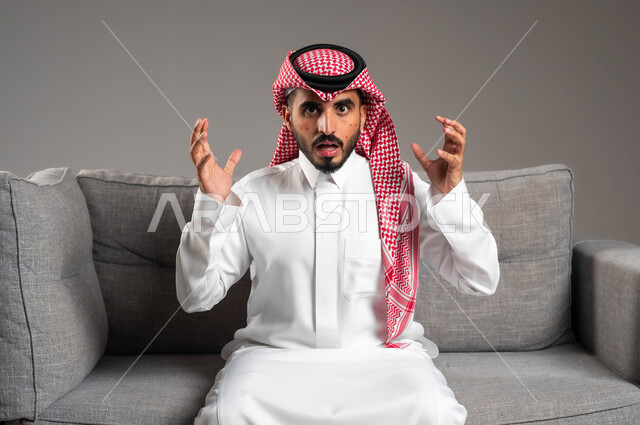 المشاكل الصحية وتهيجات الجلد, ايماءات التوتر والانزعاج وعدم تحمل الطفح الجلدي, بورتريه مقرب لشاب عربي خليجي سعودي يرتدي الثوب والشماغ التقليدي يجلس على الكنبة ويرفع يديه للأعلى, بثور منتشرة على بشرة الوجه, الإصابة الجلدية وفرط التحسس, خلفية رمادية